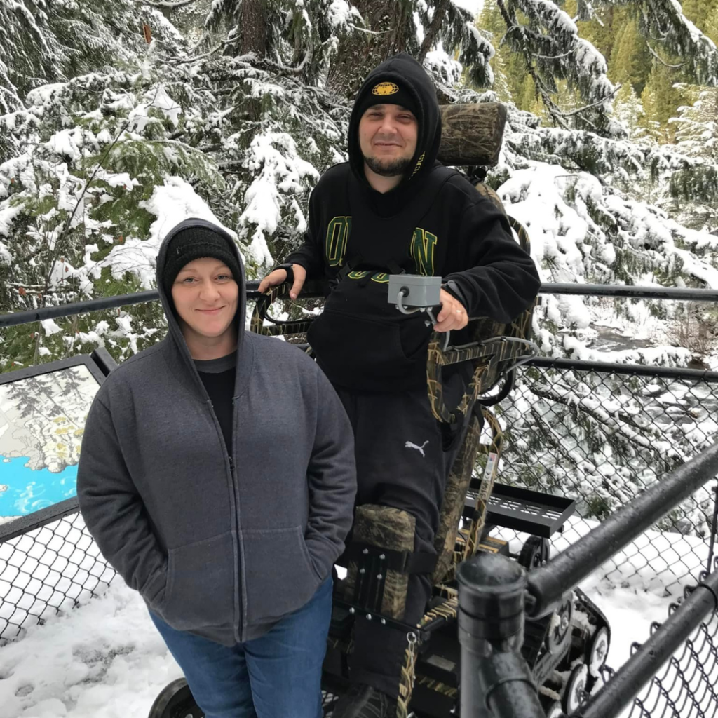 Casey in standing track chair pictured with his wife Michelle in front of snowy scenery. Casey's story of life after spinal cord injury.